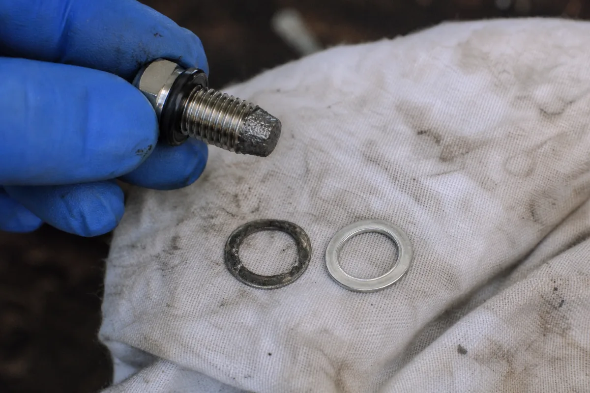 A magnetic outboard drain screw with fine gray paste on its tip, above an old crushed sealing washer and a new one.