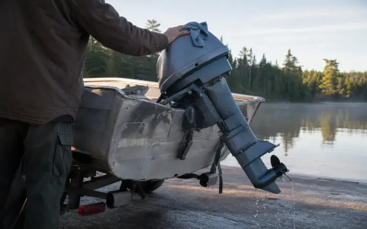 A small tiller outboard tilted up on the transom of an aluminum fishing boat at a lake boat ramp, water dripping from the propeller.