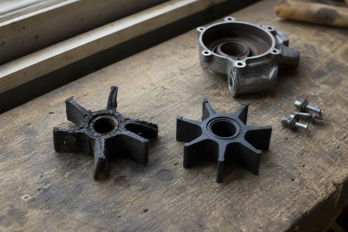 A worn outboard water pump impeller with cracked, bent vanes beside a new one on a workbench.