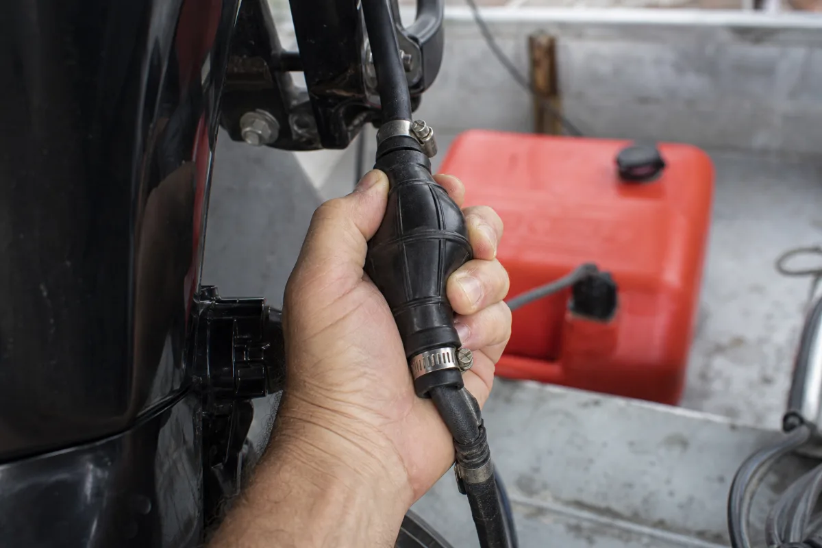 A hand squeezing the primer bulb on an outboard fuel line, with a red portable tank behind.