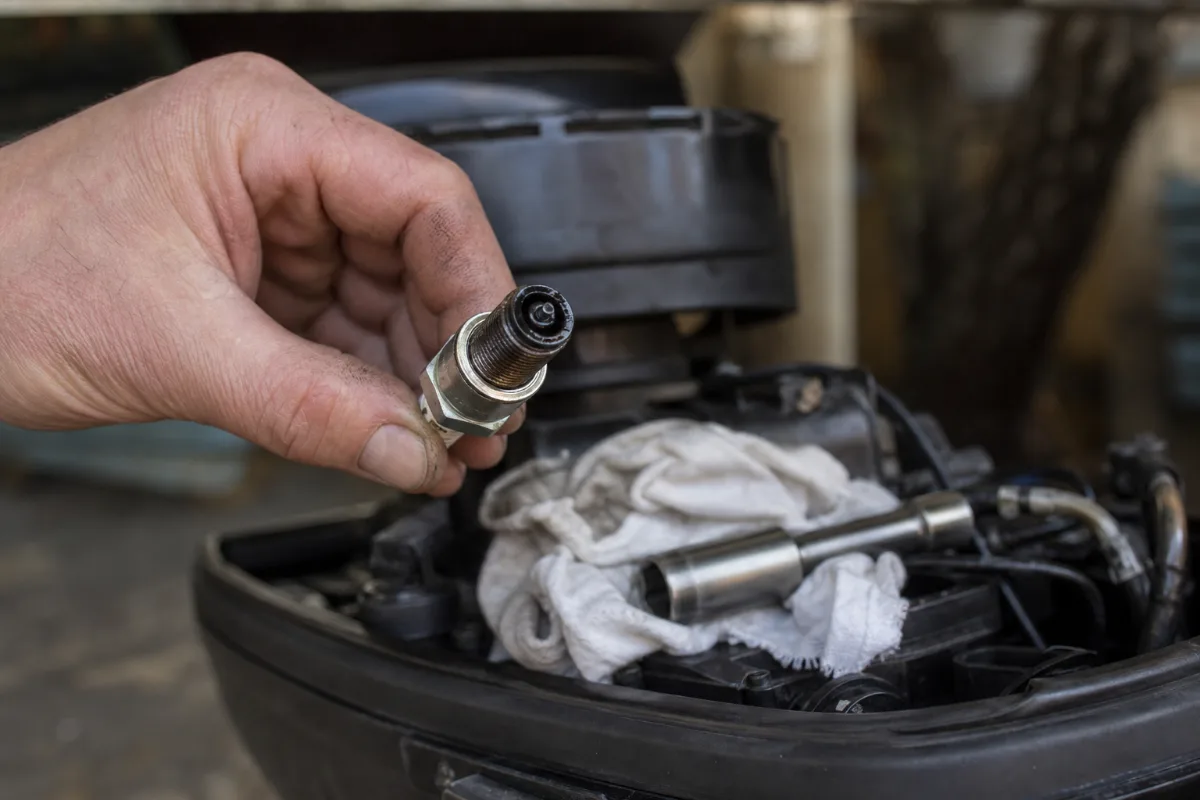 A spark plug just pulled from a small outboard, held over the engine with a plug socket and rag below.