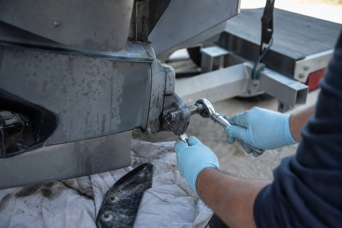 Gloved hands undoing a lower unit bolt on an outboard, with the trim tab already removed below.