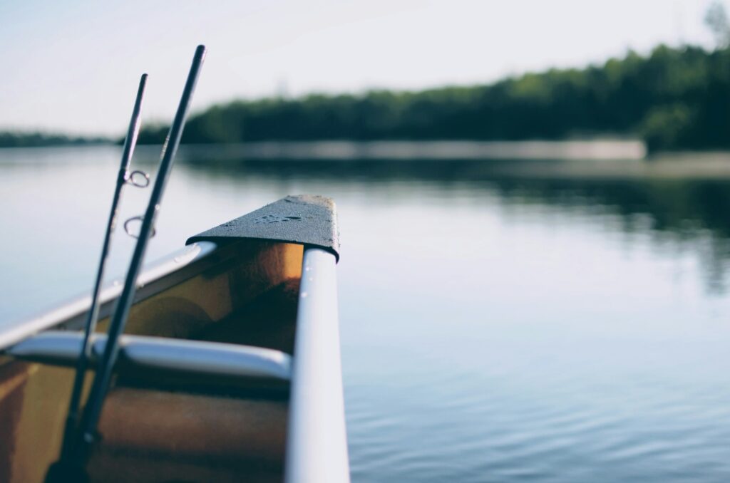 The bow of a canoe with two fishing rods standing up, a calm lake and tree line behind