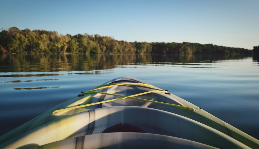 The bow of a kayak on a calm lake with a tree line beyond