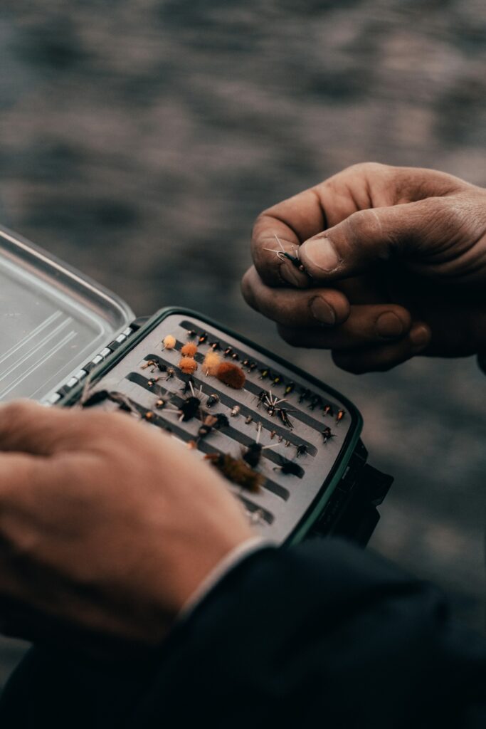 Hands picking a fly out of an open tackle box packed with rows of flies, water behind