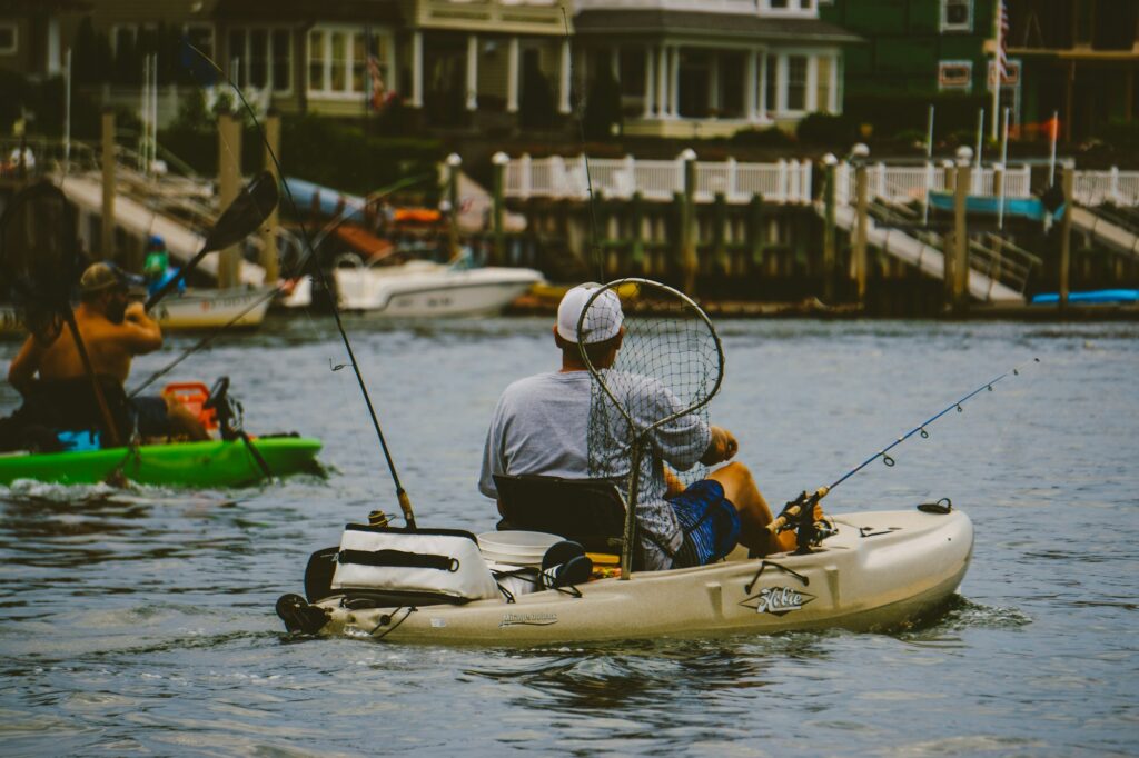 An angler in a fishing kayak with a crate, rods and a net behind the seat, another kayak nearby